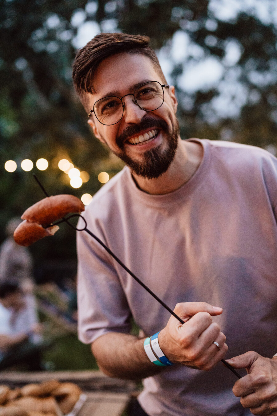 A smiling man with glasses and a beard holds a sausage skewer at an outdoor meeting, with fuzzy lights and people in the background.