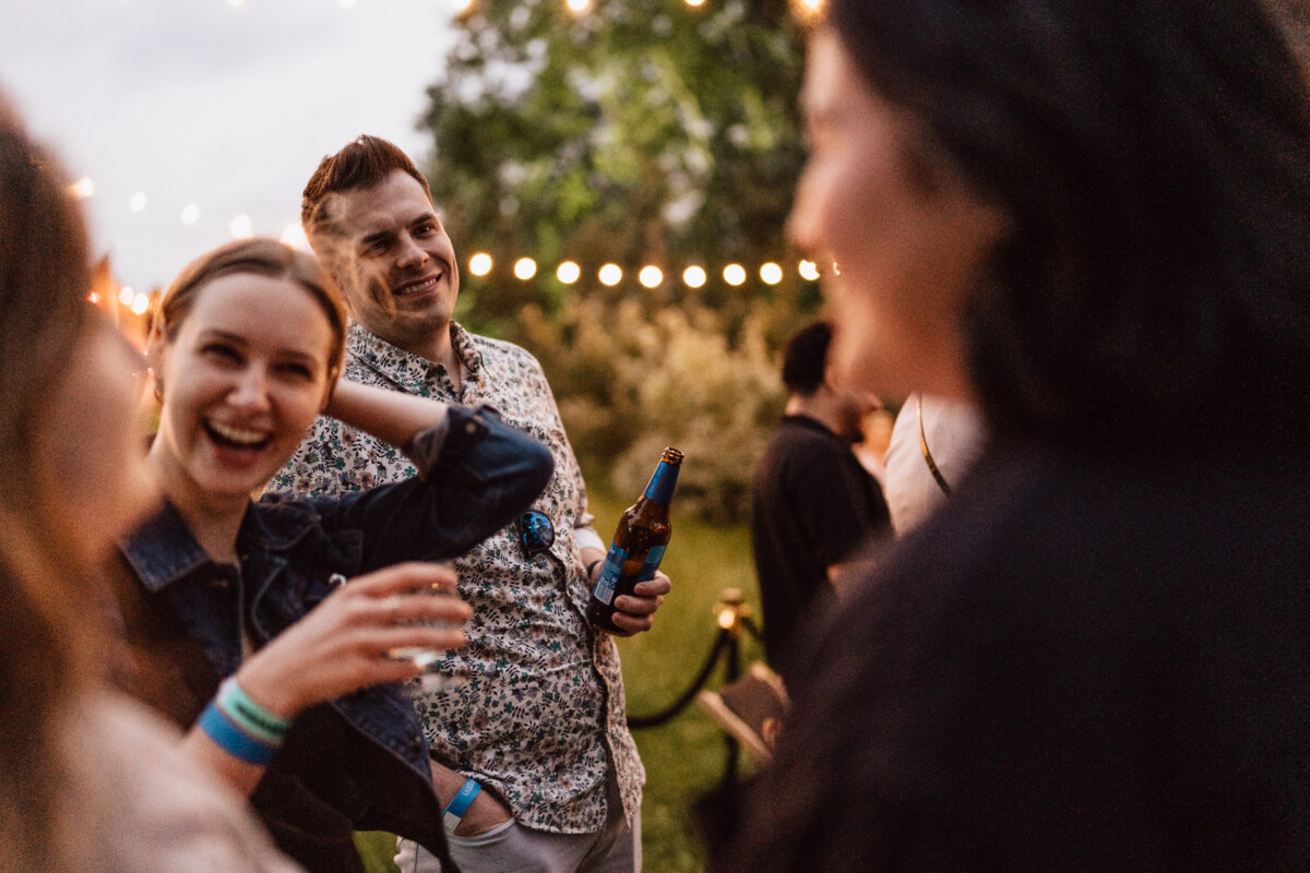 A group of people are enjoying an outdoor gathering at dusk, smiling and chatting. One person holds a bottle of beer, string lights hang above them, and trees fill the background. The overall mood is cheerful and relaxed.  