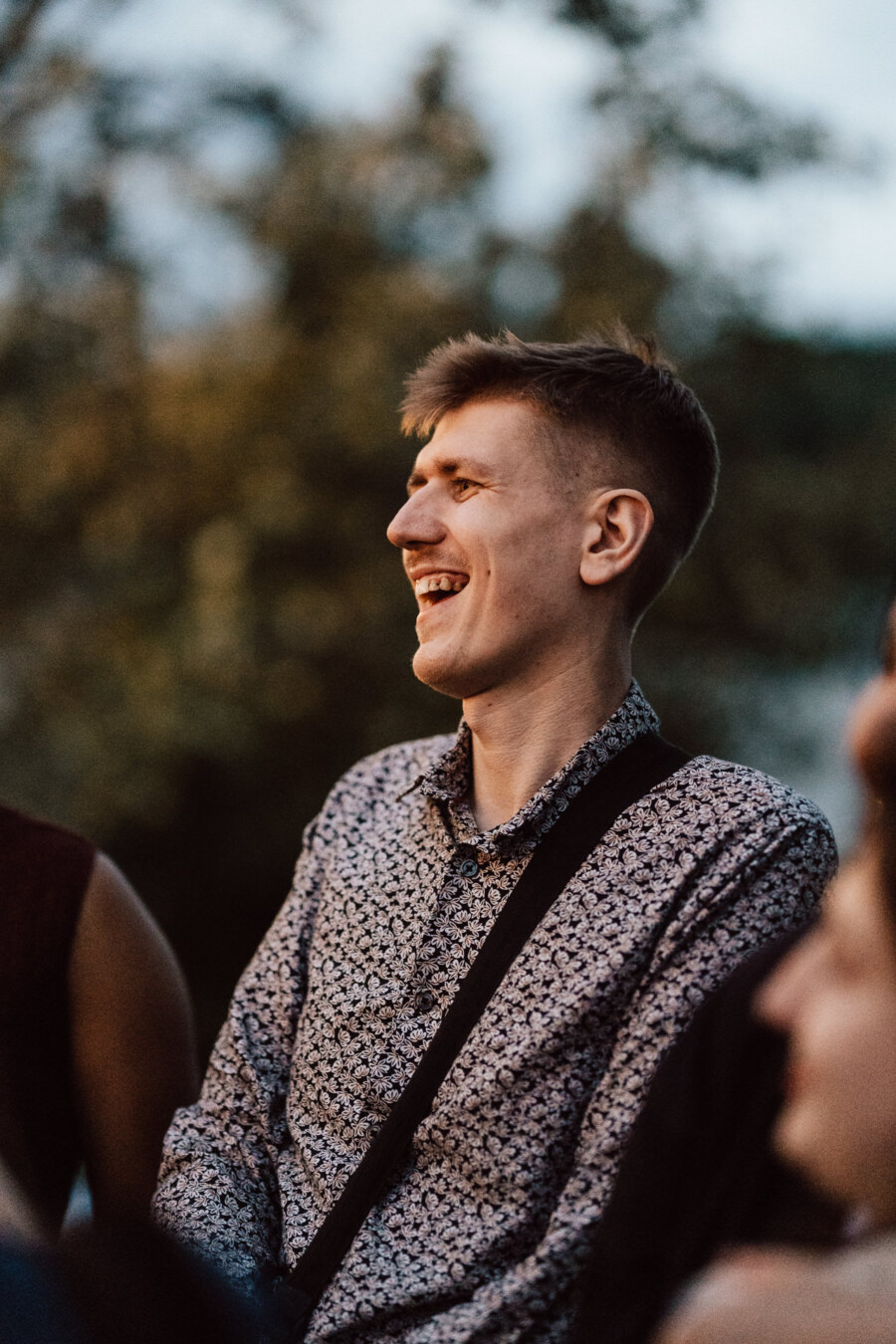 A young man with short blond hair and a patterned shirt smiles and laughs outside against a fuzzy green background.