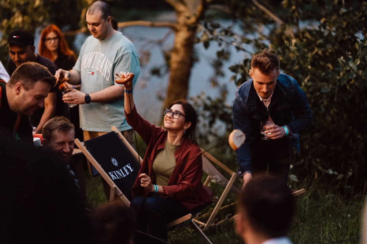 A group of people are enjoying an outdoor meeting near a river. A woman with glasses, sitting on a folding chair, reaches for food. Others stand and mingle, holding drinks, with trees and water in the background.  