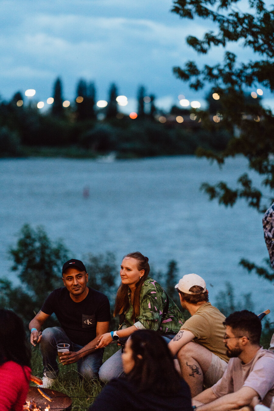 A group of people sit together outdoors by the river at dusk, talking and relaxing. Some are holding drinks, while the trees and distant city lights are visible in the background. 