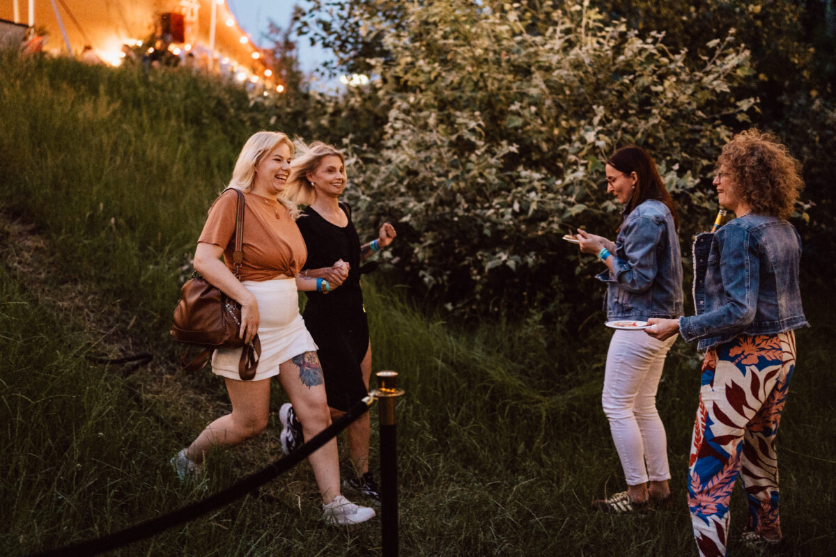 Four women smile and walk together in the evening outdoors near a grassy hill, with string lights and a tent in the background. Two walk arm in arm, while two others stand nearby, laughing and talking. 