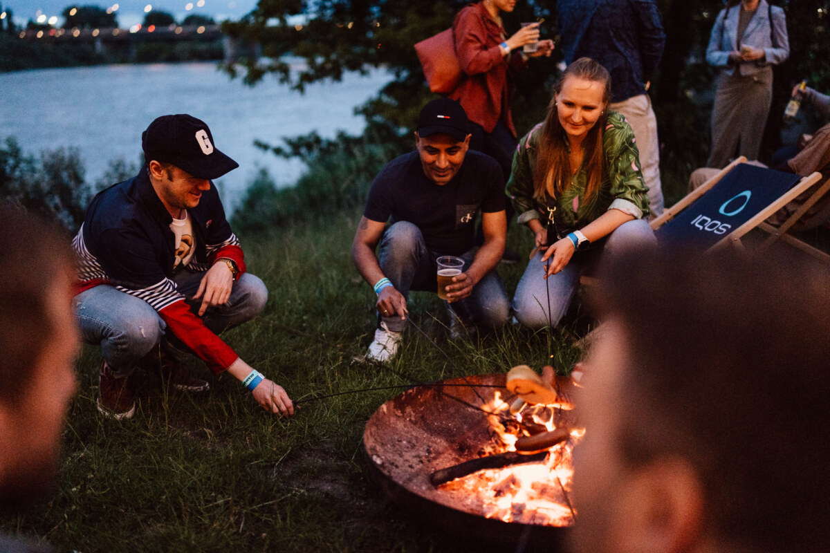 A group of people crouched around a campfire, baking food on sticks and smiling. It is evening, and they are outside near the river, surrounded by grass and trees. One person is holding a drink, and others are sitting or standing nearby.  
