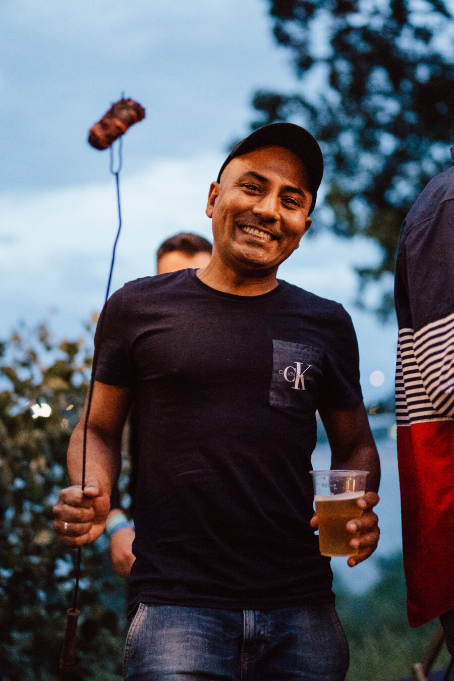 A smiling man in a dark T-shirt and cap holds a grilled sausage on a stick in one hand and a mug of beer in the other, standing outside with fuzzy trees and sky in the background.