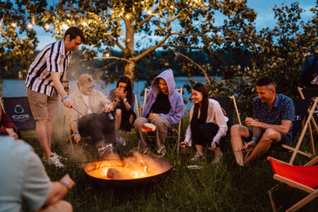 A group of people sit and stand around an outdoor campfire in the evening, baking food on sticks, laughing and enjoying each other's company under a tree with string lights.
