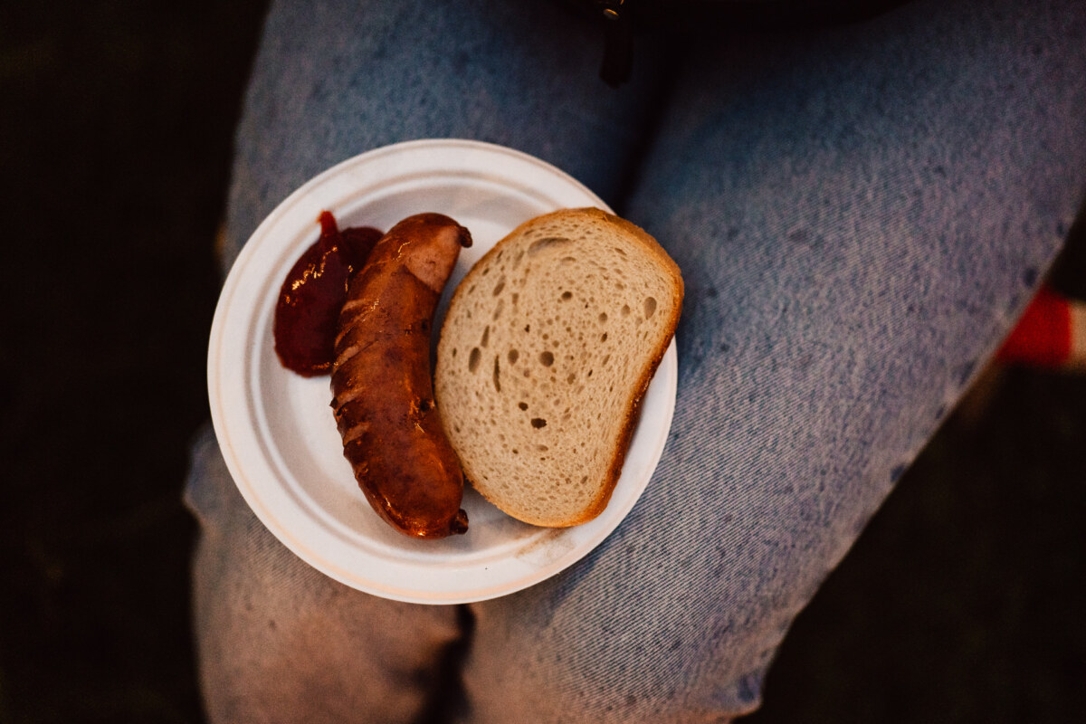 A person dressed in blue jeans holds a white paper plate with a slice of bread, grilled sausage and a portion of ketchup.