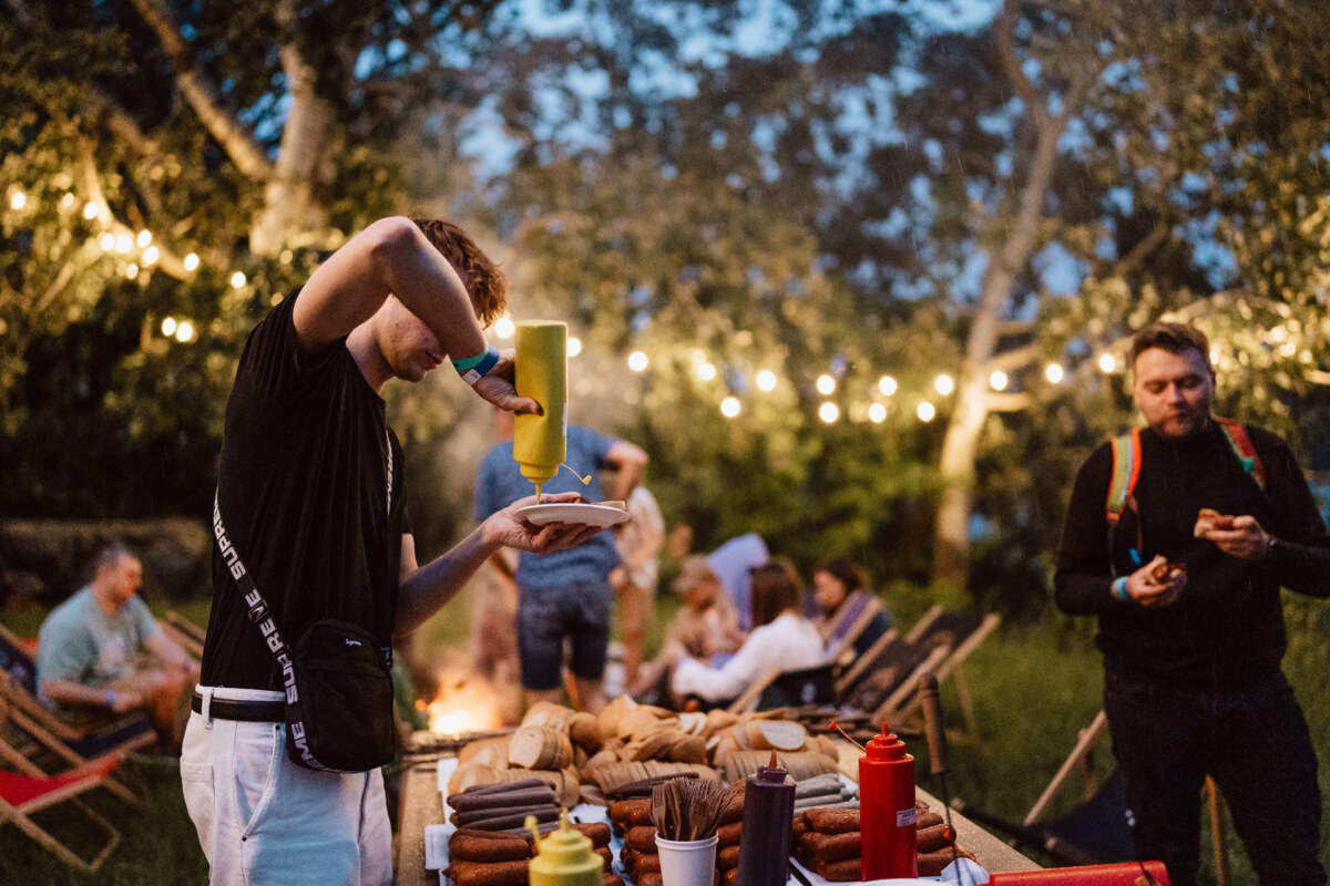 A person pours mustard on a hot dog during an outdoor gathering with several people sitting and standing under string lights in the garden. The groceries are spread out on a table in the foreground. 