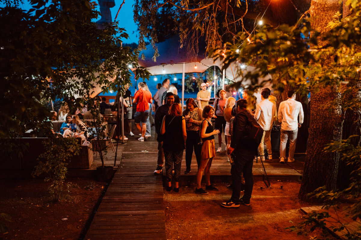 A group of people meet outdoors in the evening under string lights and trees. Some stand on the wooden sidewalk, while others sit at tables in the background, creating a lively, festive atmosphere. 