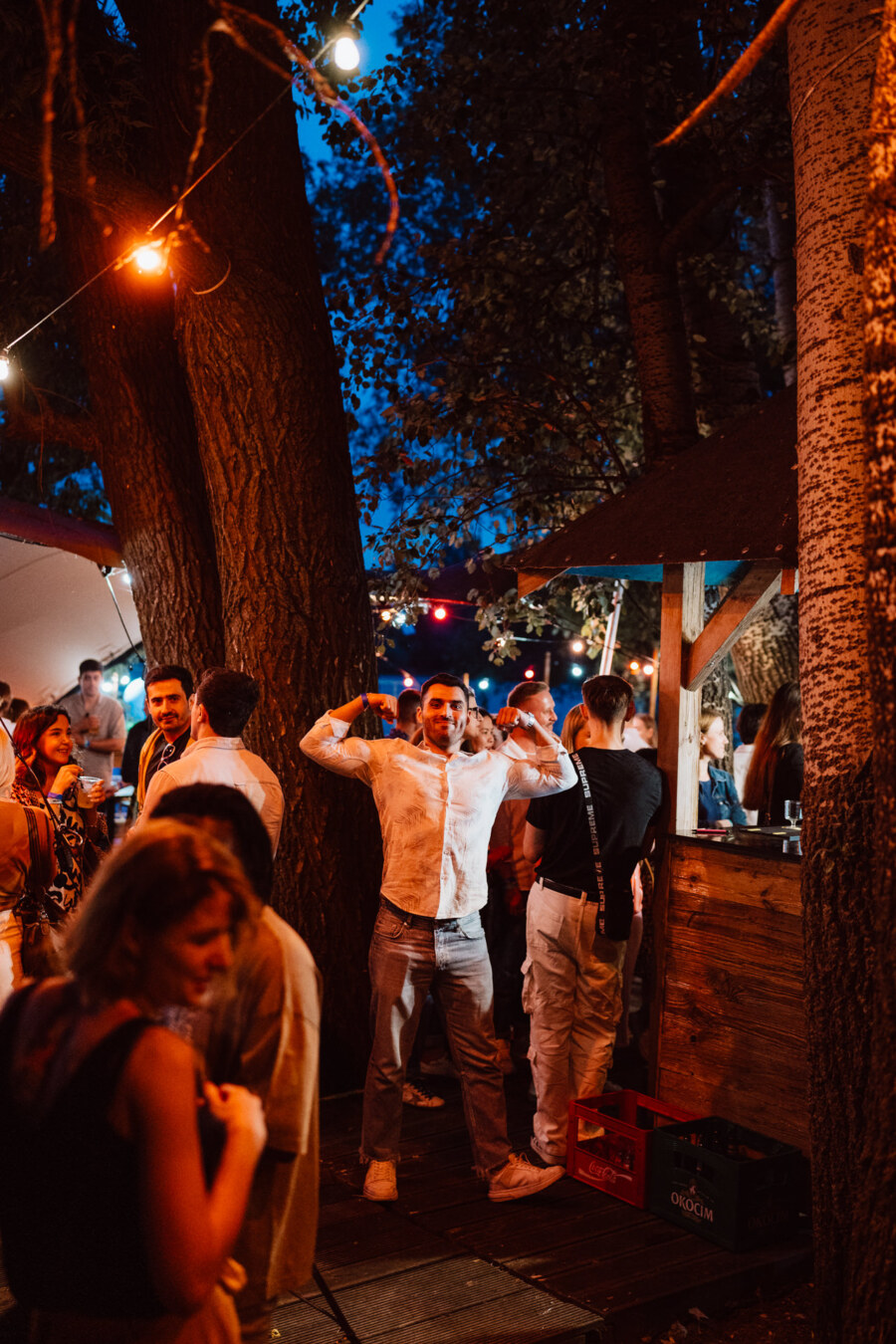 A man in a white shirt poses with his arms bent at an outdoor evening party, surrounded by people gathering under string lights and trees near a wooden bar.