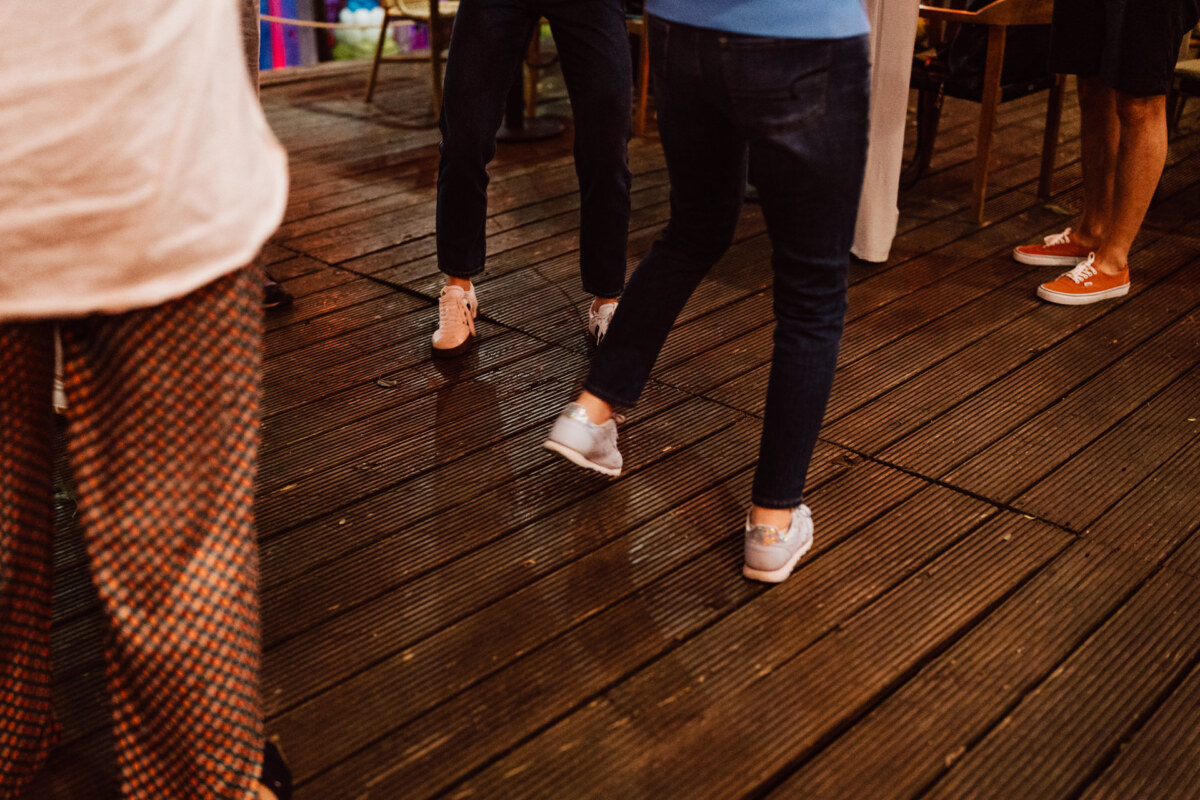 Four people are standing or walking on a wooden platform, with only their legs and feet visible. They are wearing everyday clothes and sneakers. The scenery appears to be outside, probably at an evening social event.  