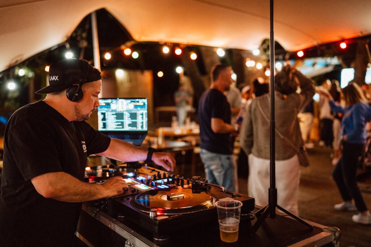 A DJ wearing headphones mixes music on a turntable during an outdoor event, accompanied by people dancing with string lights in the background. A drink is placed near the DJ's station. 