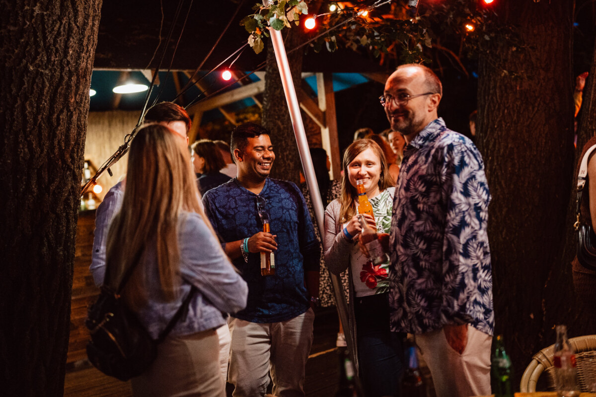 A group of people stand outdoors at night, talking and smiling under the lamps. Some are holding drinks, and they are surrounded by trees. The atmosphere is festive and relaxed.  