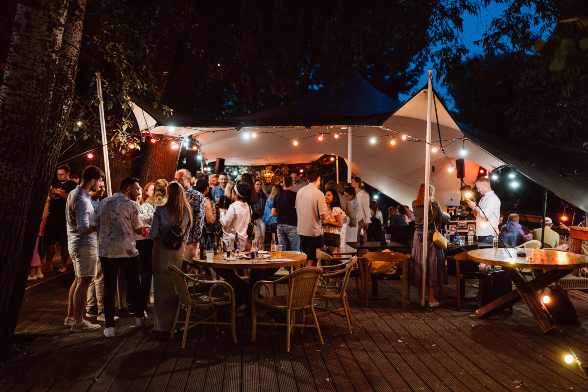 Company Party by the Vistula River - A group of people gather for an evening party outdoors under string lights and a canopy. Some talk, others stand at tables with food and drinks. Trees and warm lighting create a festive atmosphere.  