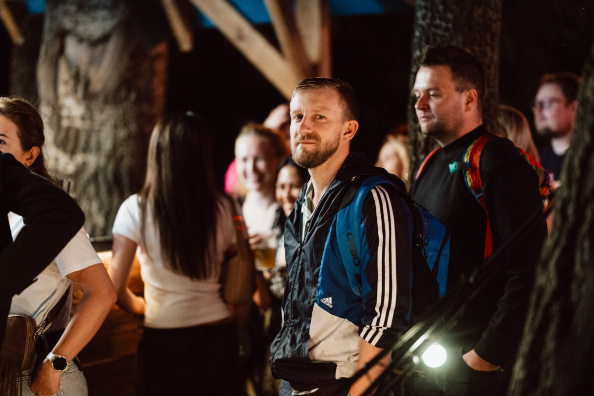 A bearded man in a blue and black jacket stands among a group of people outside at night, looking into the camera. Others around him are talking and smiling in a casual setting with trees and wooden structures. 