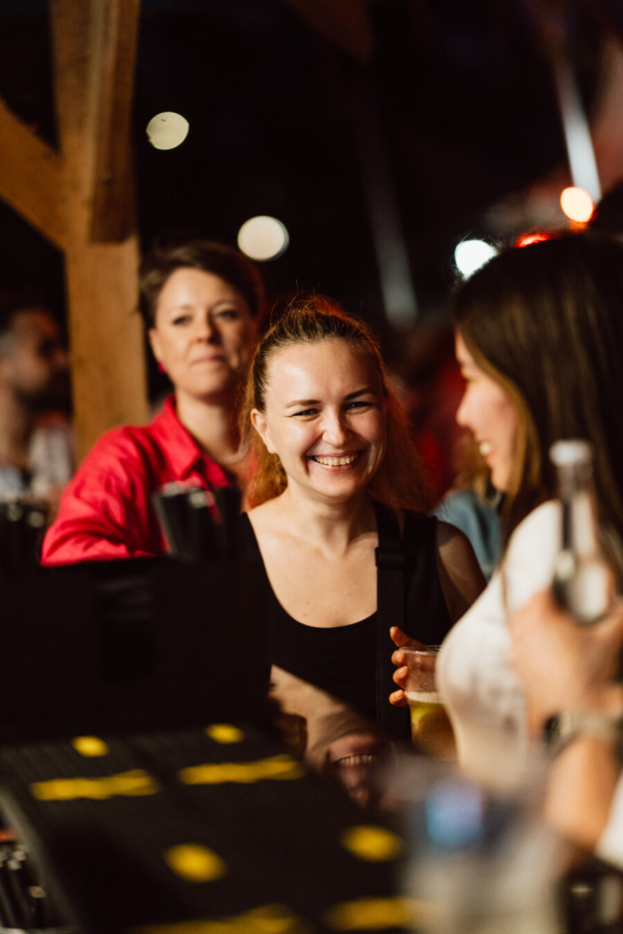 A young woman with a ponytail smiles radiantly while chatting with a friend in a dimly lit, busy bar or event space. Other people and drinks are visible in the background, creating a lively atmosphere. 