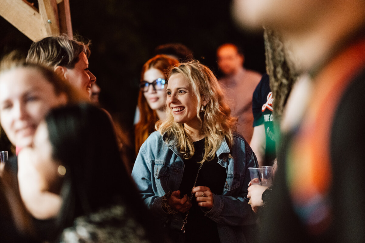A woman with blonde hair and a denim jacket smiles as she stands in a crowded open-air nightclub. Other people are gathered around her, some talking and holding drinks. 