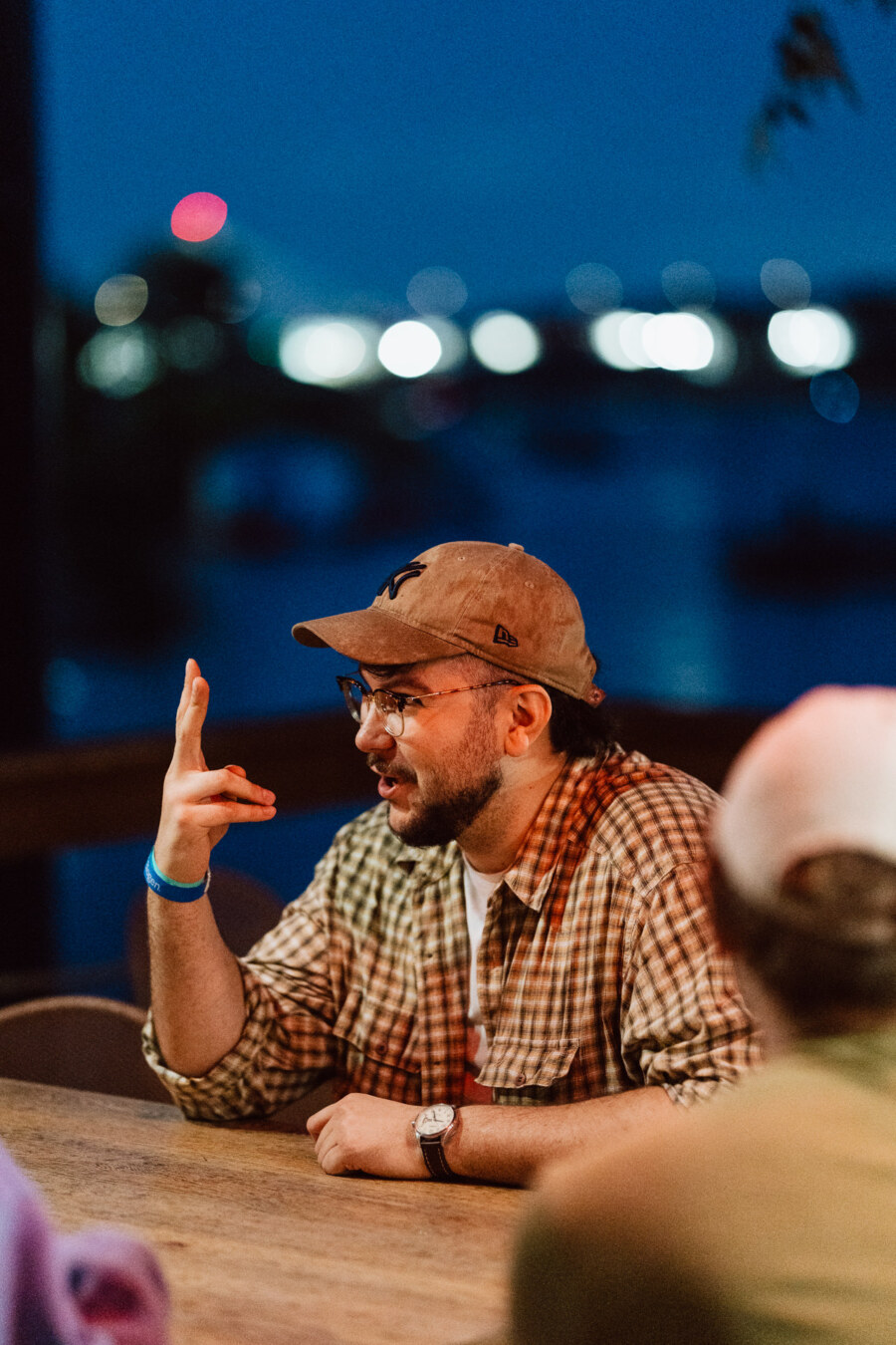 A man wearing a brown baseball cap, glasses and plaid shirt gestures with his hand while sitting at an outdoor table at dusk. Blurred lights and water are visible in the background. 