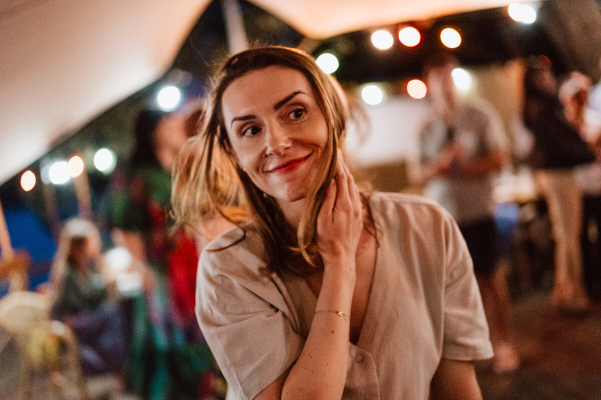 A woman with shoulder-length hair smiles as she touches her neck during a night outdoors, with fuzzy lights and people in the background.
