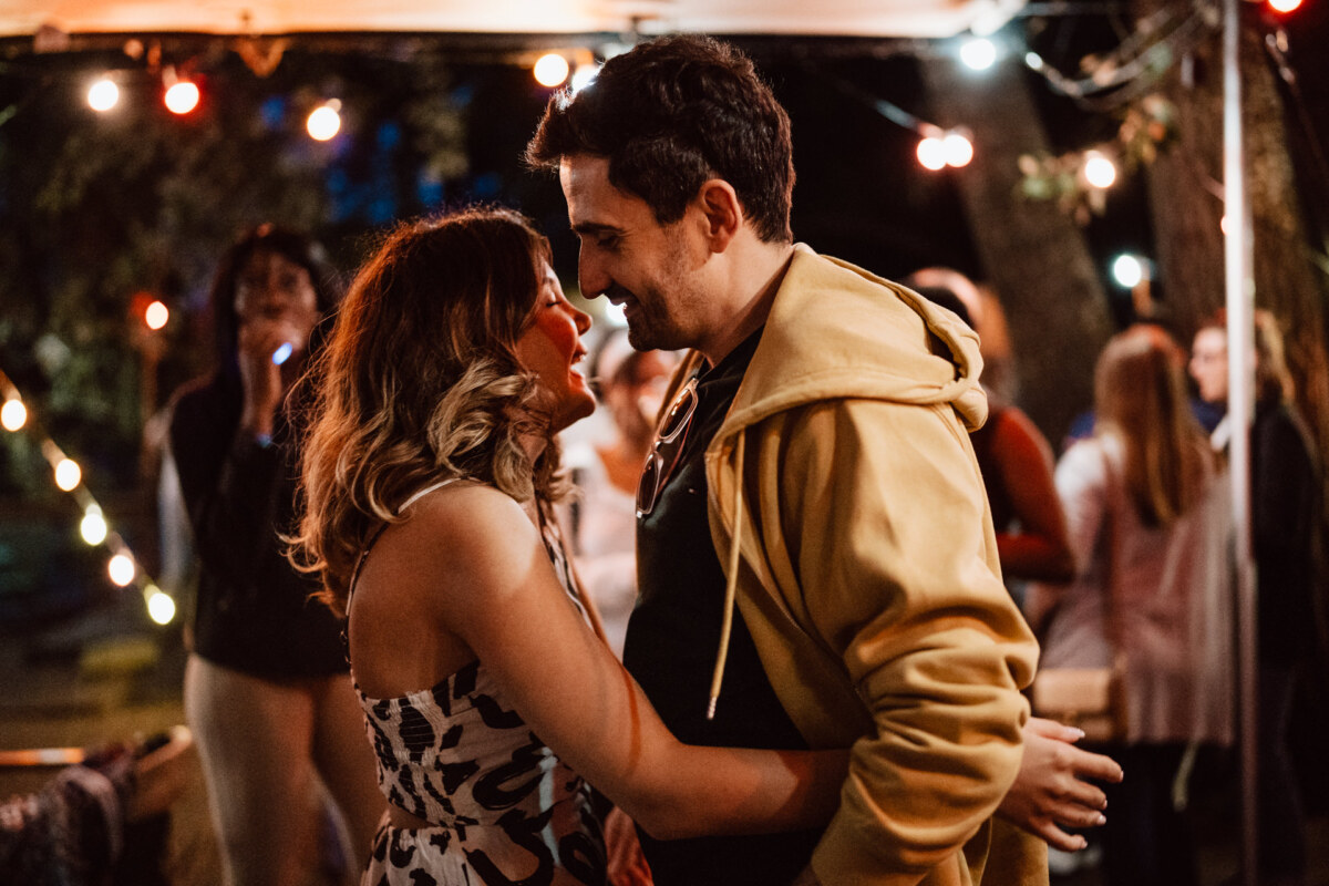 The couple embraces and smiles at each other during an outdoor evening event, surrounded by people and string lights. The atmosphere is lively and festive. 