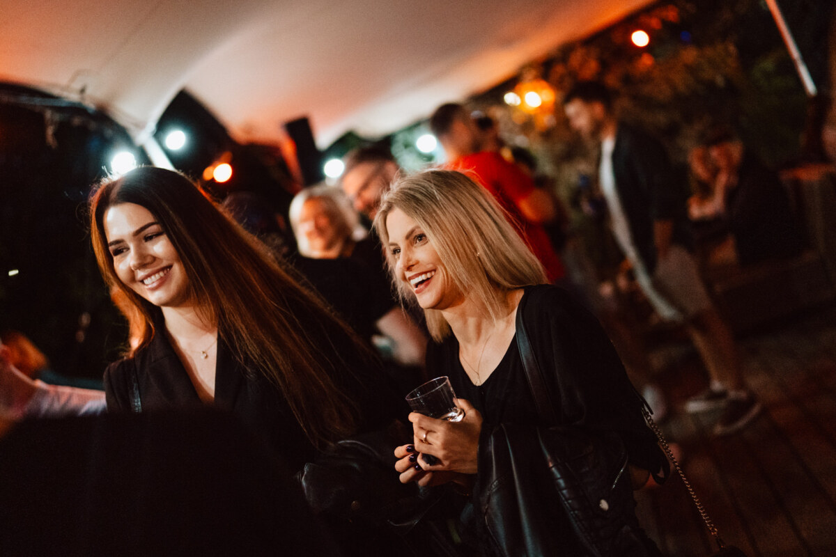 Two women smile and hold drinks at an outdoor evening party, surrounded by other people and festive lights. The atmosphere seems lively and cheerful. 