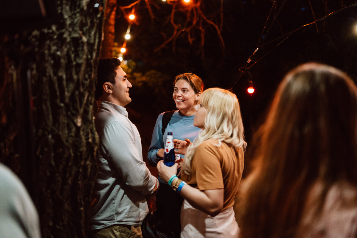 Three people stand together outside at night, smiling and talking under string lights. One person is holding a drink, and the scene is warm and festive. A fourth person with long hair is blurred in the foreground.  