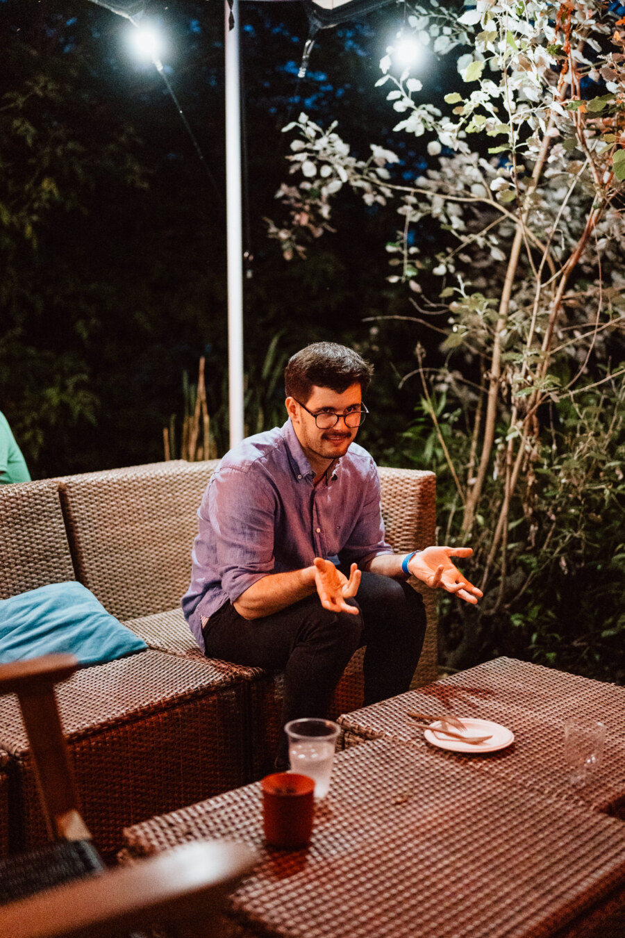 A man wearing glasses and a purple shirt sits on a wicker sofa outdoors at night, gesturing with his hands, with plates and cups on a table in front of him and trees in the background.