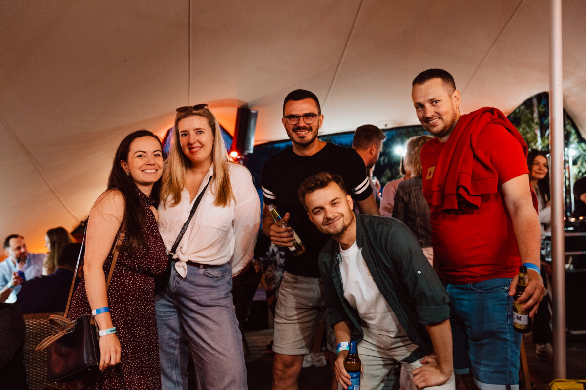 Five people pose and smile for a photo at an outdoor party, some holding drinks. They are casually dressed, in a festive and relaxed atmosphere, and other people can be seen in the background. 