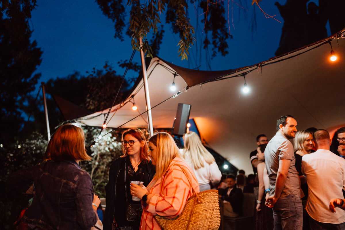 A group of people meet outdoors at night under a tent decorated with string lights. Some are talking and holding drinks while others mingle in the background. The atmosphere seems relaxed and lively.  