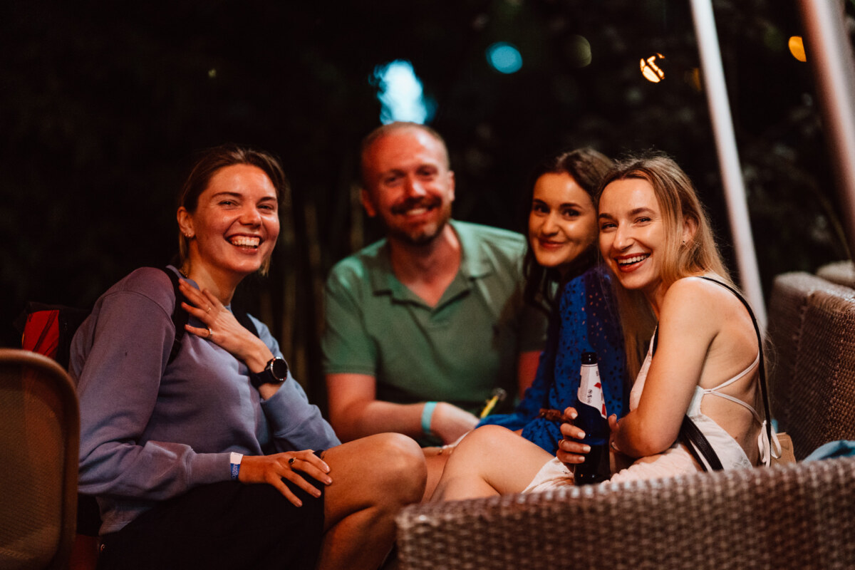 Four people sit close together on wicker furniture at night, smiling and looking happy. One of the women is holding a bottle of drink. The background is dark with dim, warm lighting.  