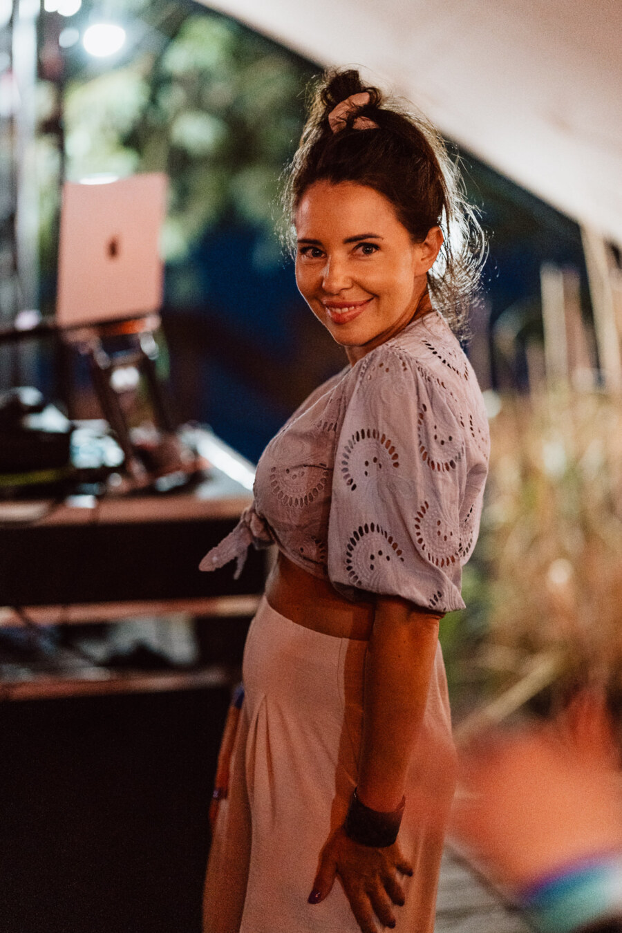 A woman with long brown hair smiles at the camera. She is wearing a light blue eyelet blouse and cream pants. The background is softly blurred, with equipment and plants visible.  