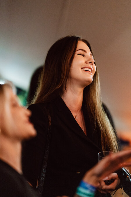 A woman with long brown hair, dressed in a black outfit, smiles and looks up during a party in the room. She looks like she's having a good time holding a drink, with blurry people in the foreground. 