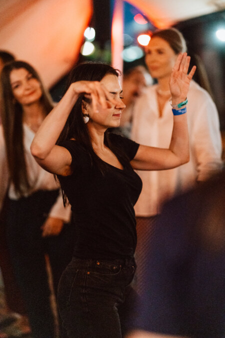 A woman in a black shirt and jeans dances with her eyes closed and hands raised, while others stand in the background at the lively, warmly lit event.