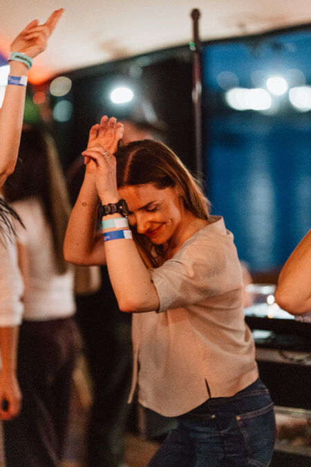 A woman with long hair dances joyfully with her hands raised at a rooftop party, wearing a beige top, blue jeans and wristbands. Blurred lights and people are visible in the background. 