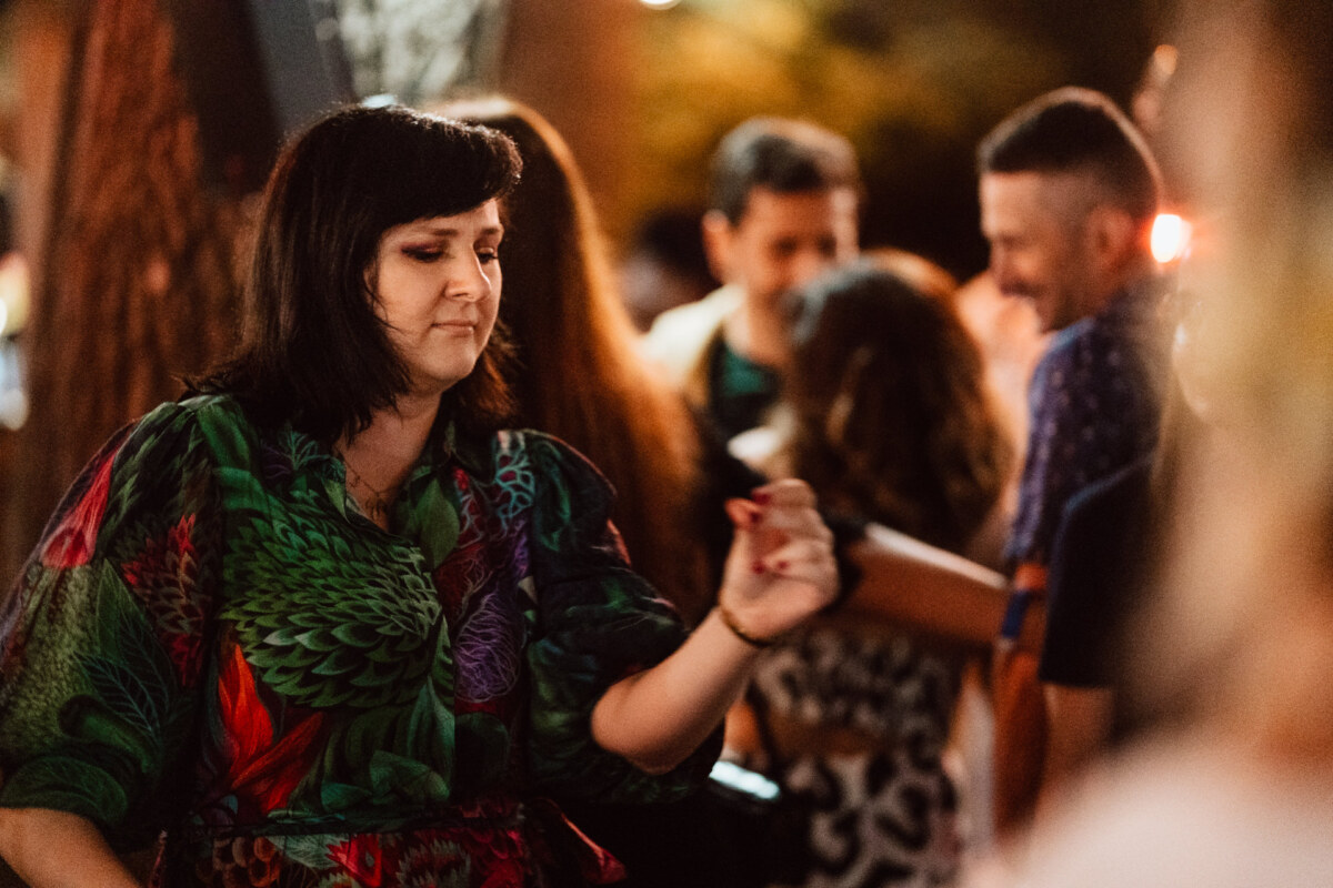 A woman in a colorful dress dances with a cheerful expression on her face during a night party outdoors, while blurry people gather in the background.