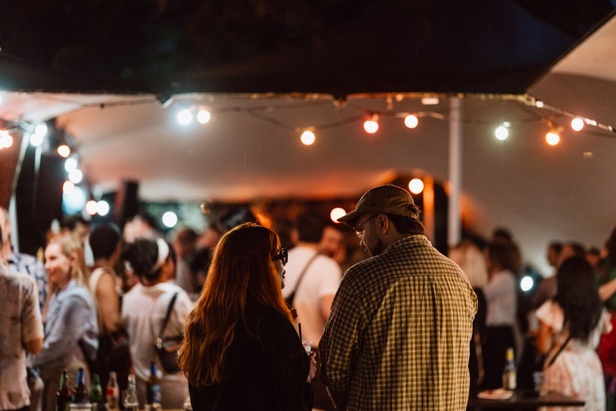 A crowd of people meet at an outdoor party at night by string lights. In the foreground, a man and woman stand face to face, engaged in conversation, while others mingle in the background. 