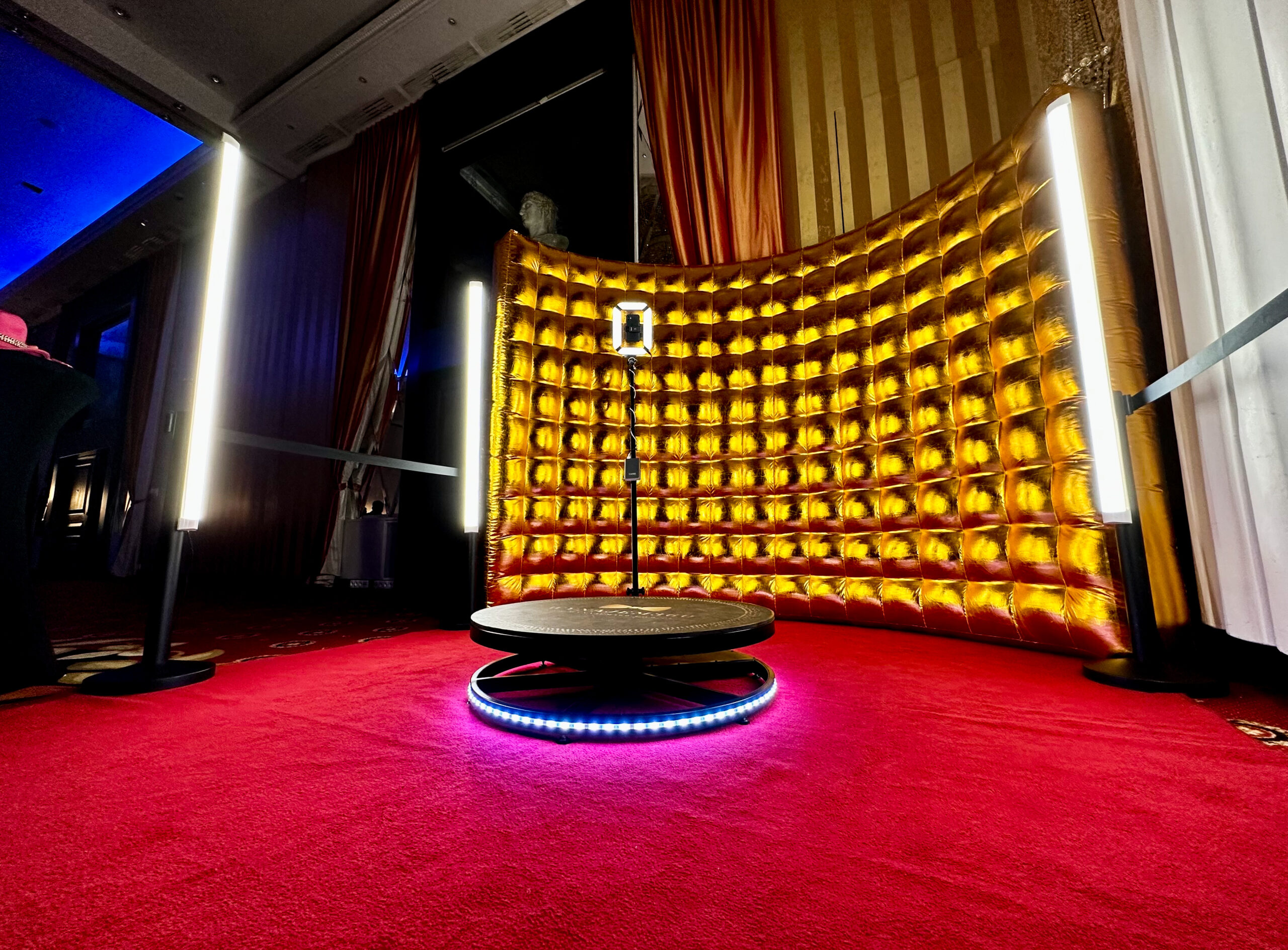 Zdjecie w tle scaled 1 Budka fotograficzna z okrągłą platformą i światłem pierścieniowym stoi na czerwonym dywanie przed złotym, pikowanym tłem, otoczonym dwoma pionowymi światłami LED w słabo oświetlonym pomieszczeniu.