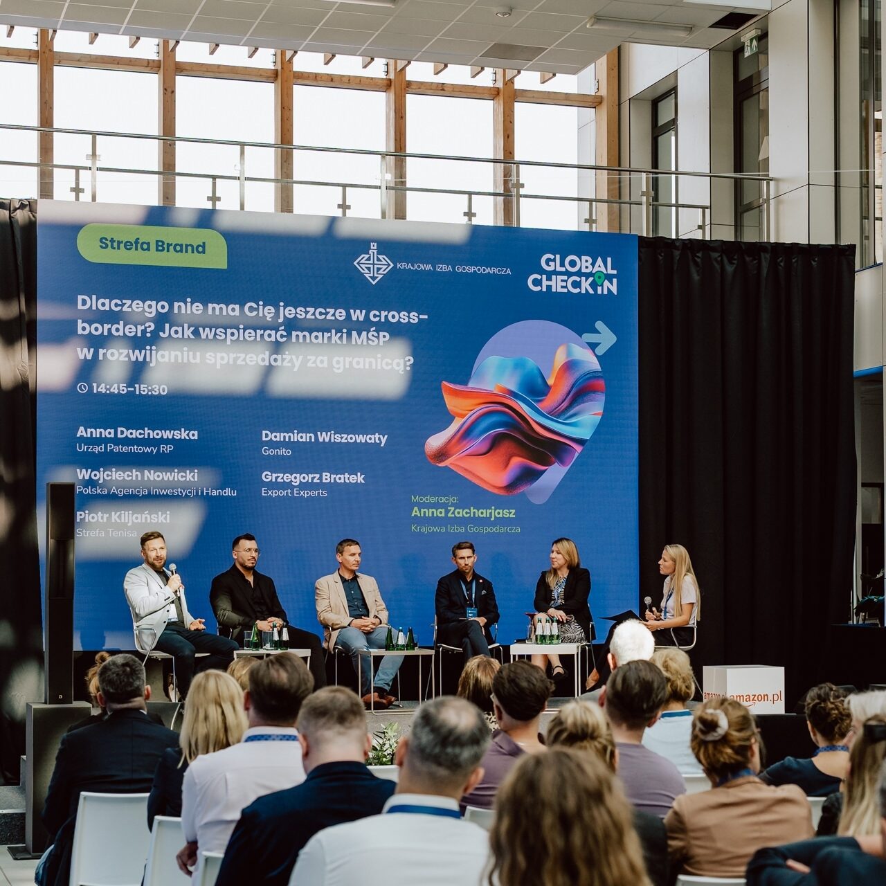 fotograf na konferencje global Check in 085 edited Eine Gruppe von sechs Rednern sitzt auf der Bühne vor einem großen blauen Hintergrund, auf dem Text und bunte Grafiken zu sehen sind. Das Publikum sitzt vorne und hört zu. Der Raum hat große Fenster und ein modernes Design.