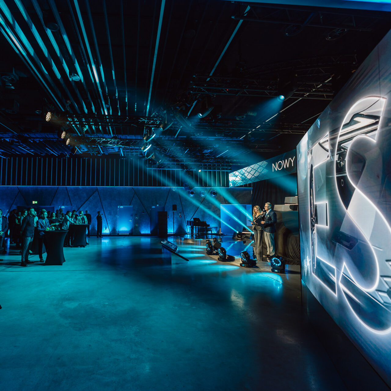 fotografia eventowa premiera lexus es200 warszawa koneser 026 edited Ein moderner, geräumiger Veranstaltungsraum, der mit blauem Licht beleuchtet wird, mit einer Bühne mit Moderatoren, einem Publikum an Cocktailtischen und einem großen beleuchteten 'ES'-Schild auf der rechten Seite.