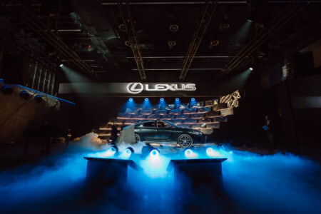Fotografia Eventowa Premium - Srebrny samochód Lexus jest wyświetlany na scenie otoczonej niebieskimi światłami i mgłą, z dużym podświetlanym logo Lexusa powyżej i nowoczesnym metalicznym wystrojem w tle.