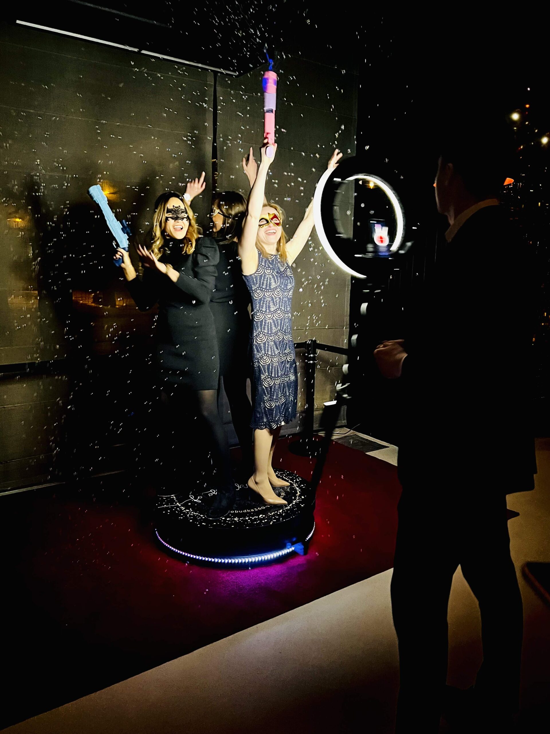 zdjecie dodatkowe do wykorzystania na stronie3 scaled 1 Trzy kobiety w imprezowych sukienkach i okularach przeciwsłonecznych pozują figlarnie na oświetlonej platformie z opadającym konfetti, podczas gdy mężczyzna filmuje je za pomocą smartfona na statywie z lampą pierścieniową w ciemnym wnętrzu.