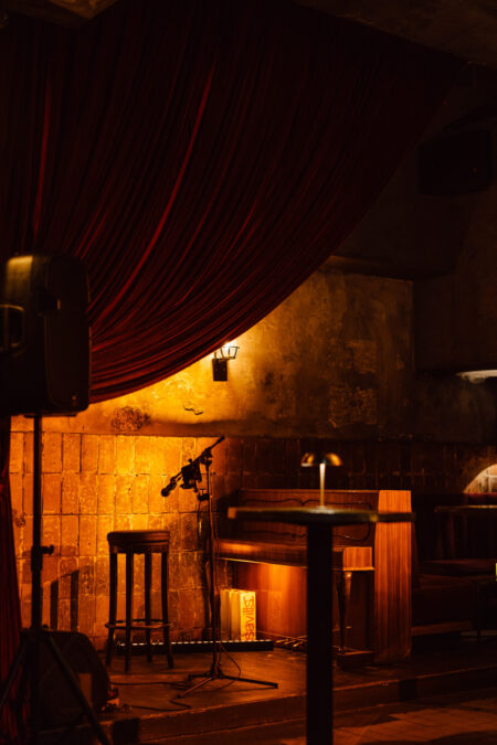 Fotograf na impreze firmowa weles bar warszawa1 Słabo oświetlona scena w przytulnym barze zawiera mikrofon, stołek i stojak na nuty. Ciemnoczerwone zasłony i ciepłe żółte oświetlenie tworzą intymną atmosferę z drewnianymi ławkami i odsłoniętymi ceglanymi ścianami w tle.