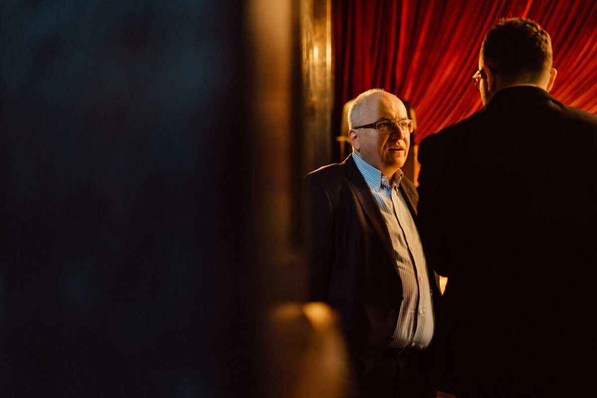 Fotograf na impreze firmowa weles bar warszawa10 1 Mężczyzna w garniturze i okularach stoi w pomieszczeniu, częściowo oświetlony ciepłym światłem, rozmawiając z innym mężczyzną plecami do kamery. W tle wiszą czerwone zasłony, tworząc intymną atmosferę.