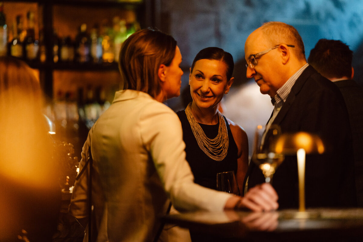 Fotograf na impreze firmowa weles bar warszawa12 Trzy osoby stoją razem w słabo oświetlonym barze, zaangażowane w rozmowę. Jedna kobieta uśmiecha się, druga stoi tyłem do kamery, a mężczyzna w okularach słucha. W tle widoczne są butelki.