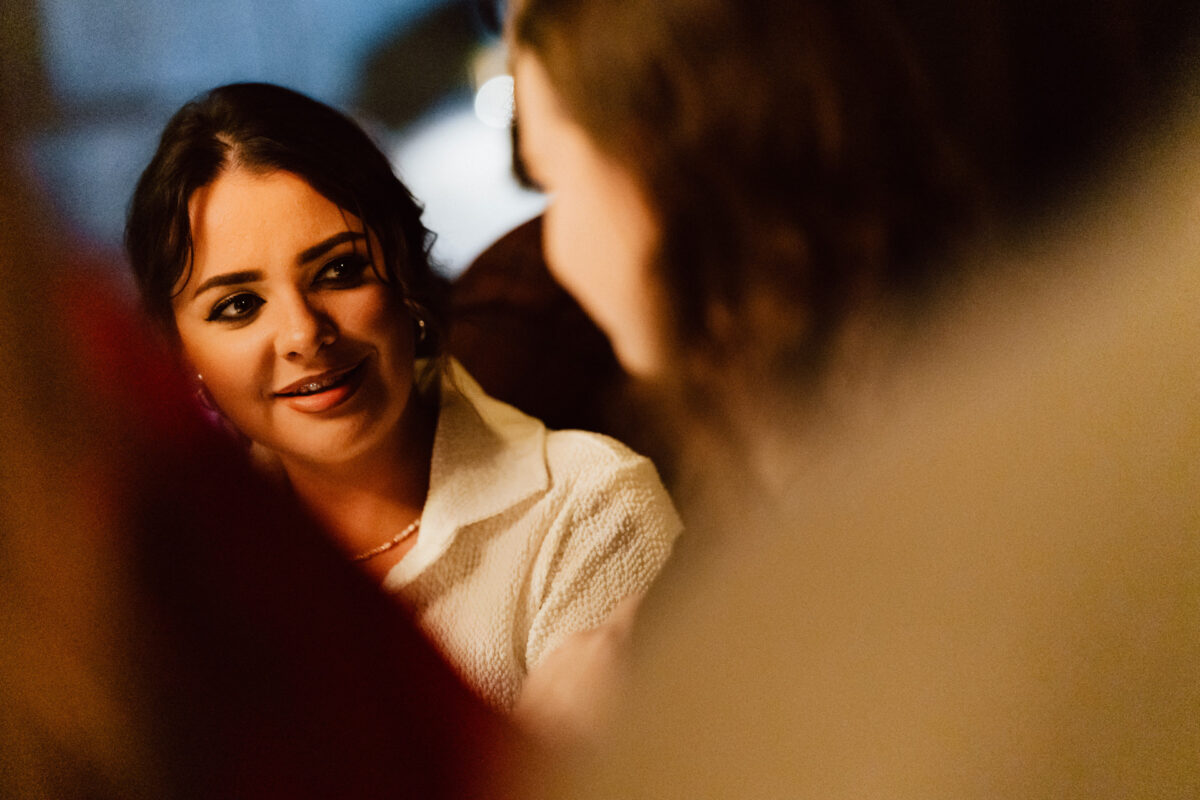 Fotograf na impreze firmowa weles bar warszawa13 Kobieta z ciemnymi włosami i białym topem uśmiecha się ciepło podczas rozmowy z kimś w delikatnie oświetlonym, przytulnym otoczeniu. Ostrość skupia się na jej twarzy, podczas gdy osoba, z którą rozmawia, jest rozmyta na pierwszym planie.