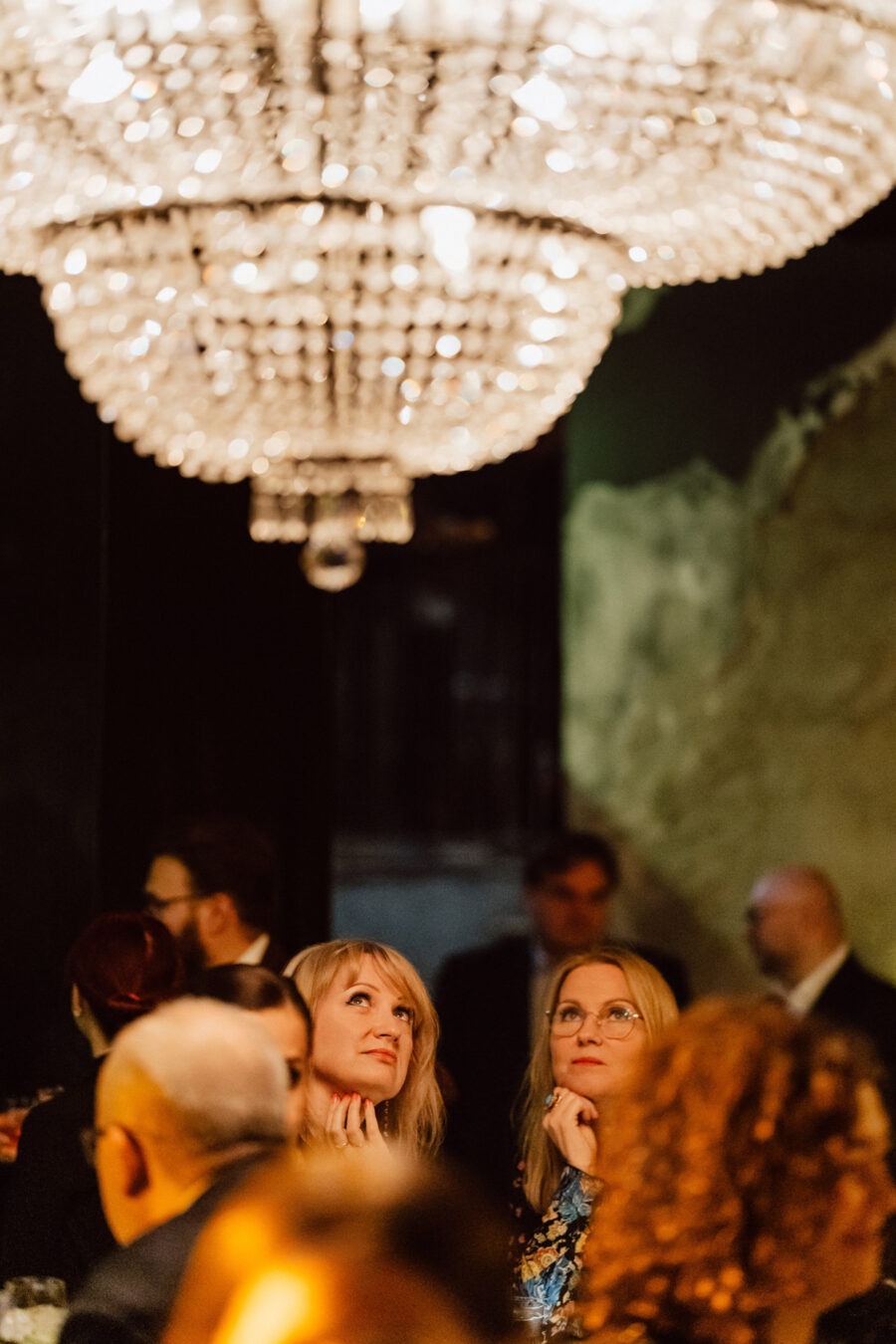 Fotograf na impreze firmowa weles bar warszawa14 Dwie kobiety siedzą blisko siebie, patrząc w zamyśleniu na duży, ozdobny żyrandol nad głową. Inni ludzie są widoczni w delikatnie oświetlonym, eleganckim pokoju w tle.