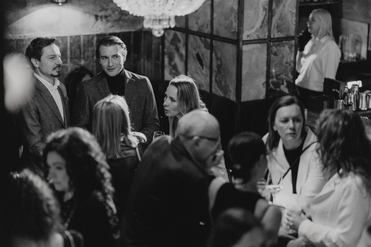 Fotograf na impreze firmowa weles bar warszawa15 Czarno-białe zdjęcie zatłoczonego baru z ludźmi siedzącymi i stojącymi, zaangażowanymi w rozmowę. Dwóch mężczyzn stoi i rozmawia z kobietą, podczas gdy inni siedzą przy stolikach. W tle widoczny jest barman.