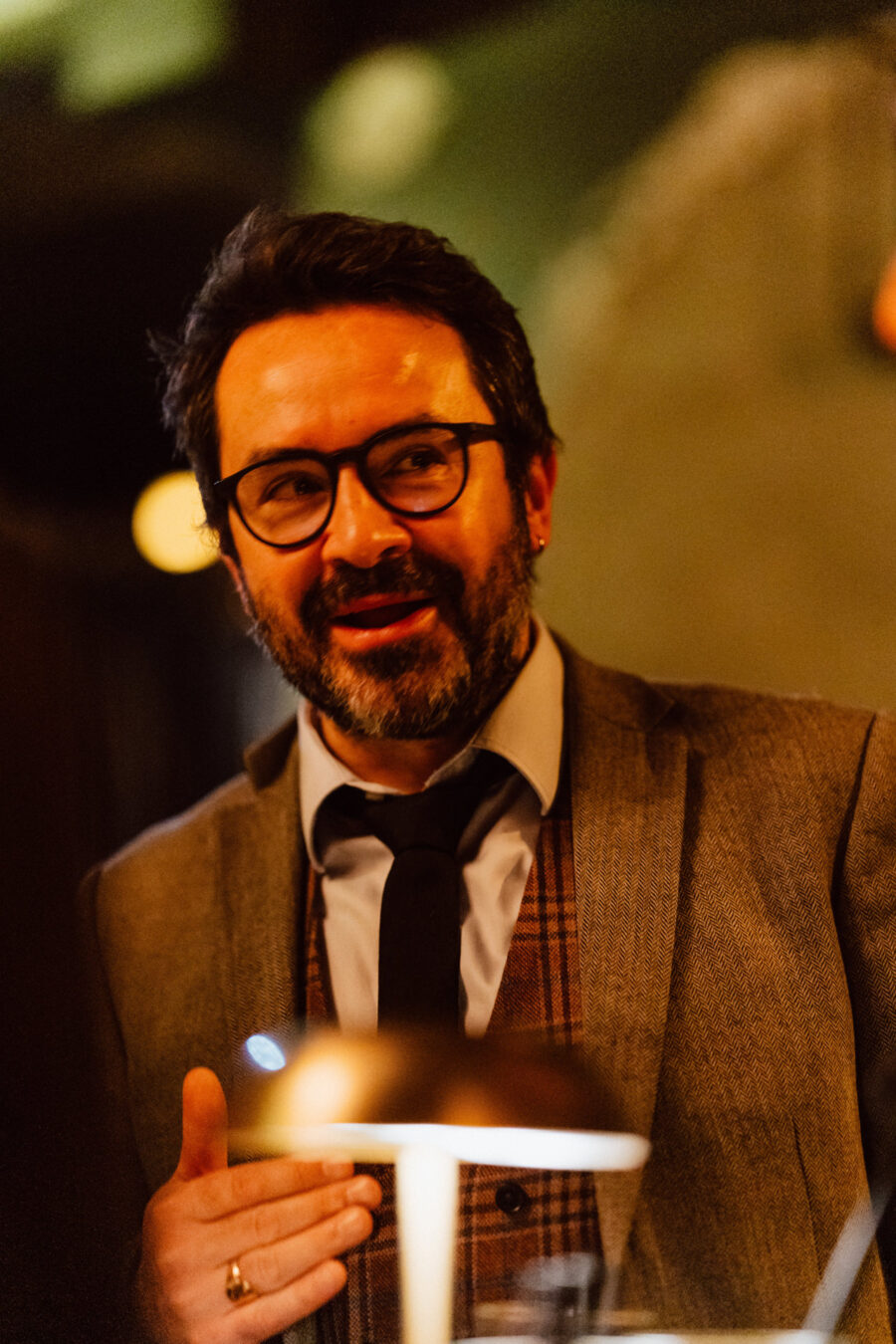 Fotograf na impreze firmowa weles bar warszawa17 Mężczyzna w okularach, z ciemną brodą, ubrany w garnitur i krawat, gestykuluje podczas rozmowy w ciepło oświetlonym pomieszczeniu.