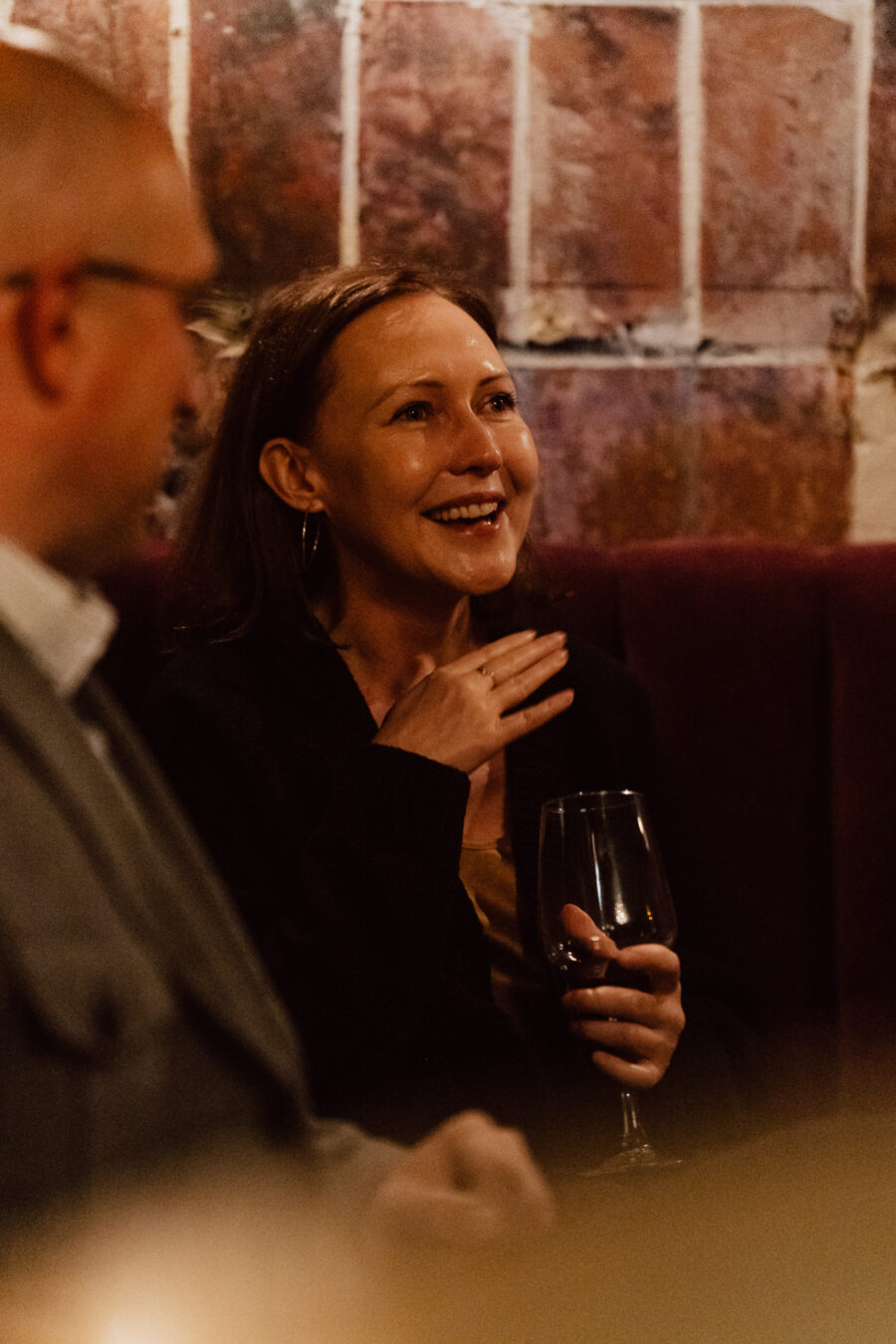 Fotograf na impreze firmowa weles bar warszawa18 Uśmiechnięta kobieta z kieliszkiem czerwonego wina siedzi w słabo oświetlonej restauracji i wydaje się być zaangażowana w rozmowę z mężczyzną obok niej. Tło stanowią odsłonięte ceglane ściany.
