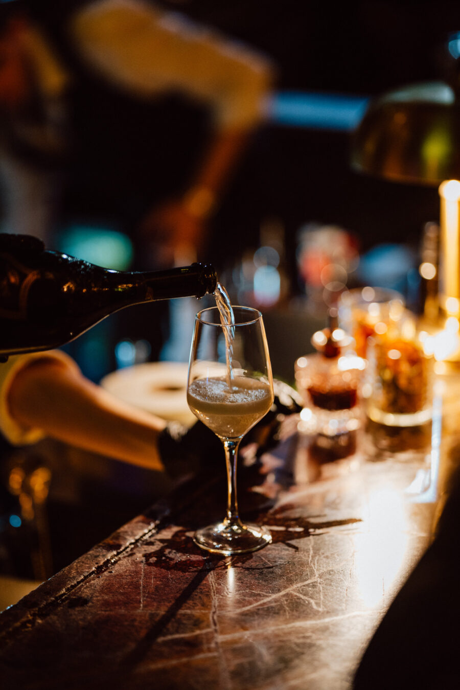 Fotograf na impreze firmowa weles bar warszawa19 Zbliżenie ręki nalewającej białe wino z butelki do kieliszka na słabo oświetlonej ladzie barowej, z butelkami i słoikami w rozmytym tle.