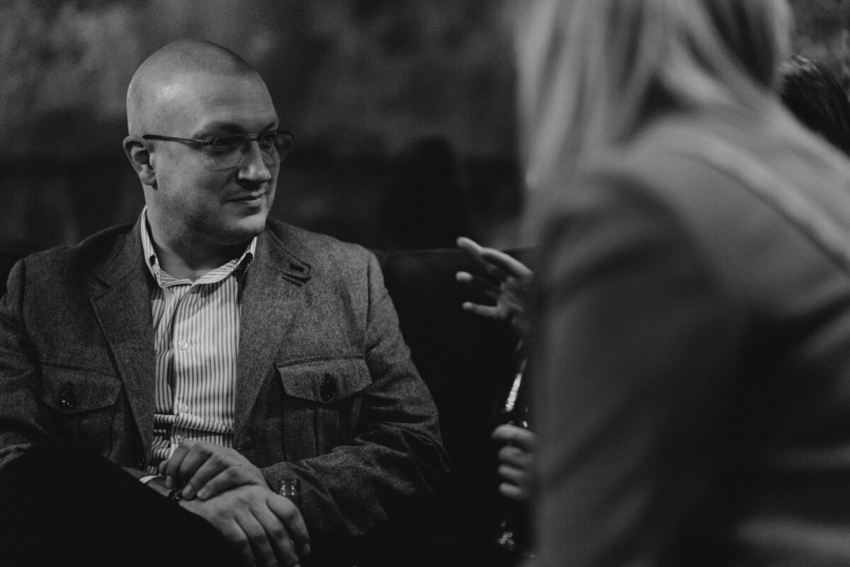 Fotograf na impreze firmowa weles bar warszawa20 1 Mężczyzna w okularach i marynarce siedzi, słuchając uważnie osoby na pierwszym planie, która gestykuluje ręką. Obraz jest czarno-biały i ma swobodną, konwersacyjną atmosferę.